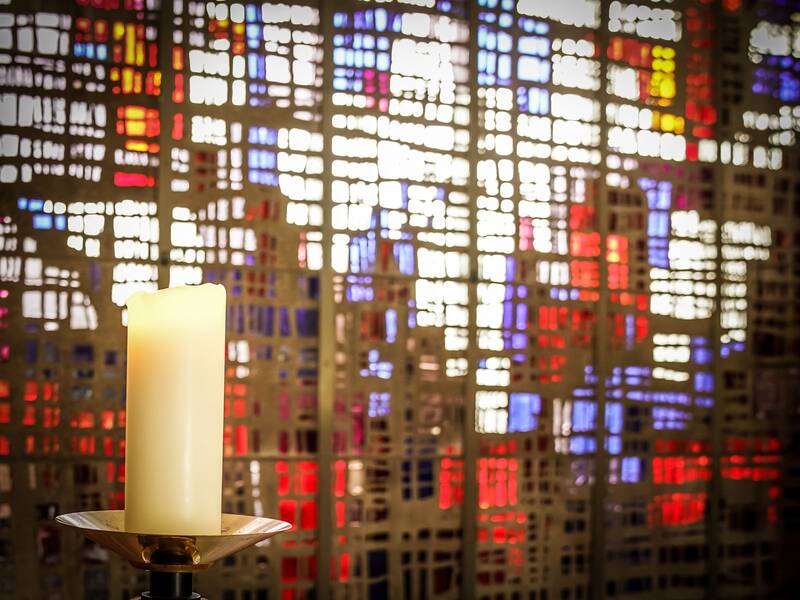 Bild aus der Aussegnungshalle Aldingen. Im Vordergrund eine brennende Kerze im Hintergund die bunte Bleiverglasung