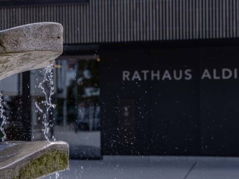 Ein Steinbrunnen sprudelt Wasser vor einem modernen Gebäude mit der Aufschrift "Rathaus Aldingen".