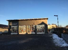 Bild zeigt eine große Garage mit 4 durchsichtigen Toren, hinter denen Feuerwehrfahrzeuge zu sehen sind.